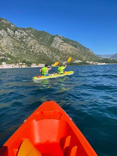 View from the front of a kayak, following another kayak with two people paddling on the calm blue sea, with the green coastline in the distance.