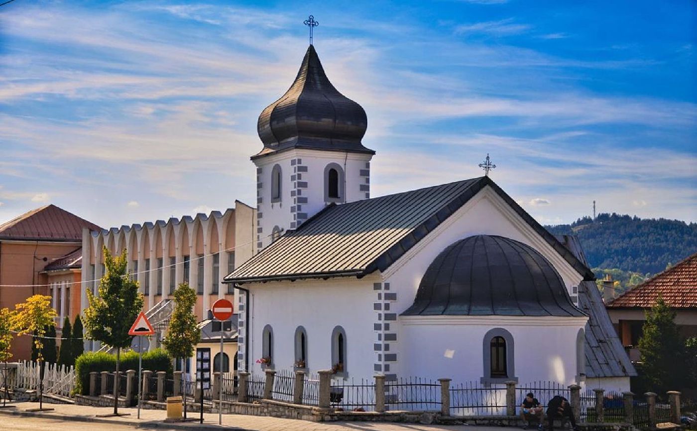 The white Orthodox Church of St. Petka in Pljevlja, Montenegro, featuring a distinctive black onion dome, grey stone accents, and a modern building in the background under a blue sky