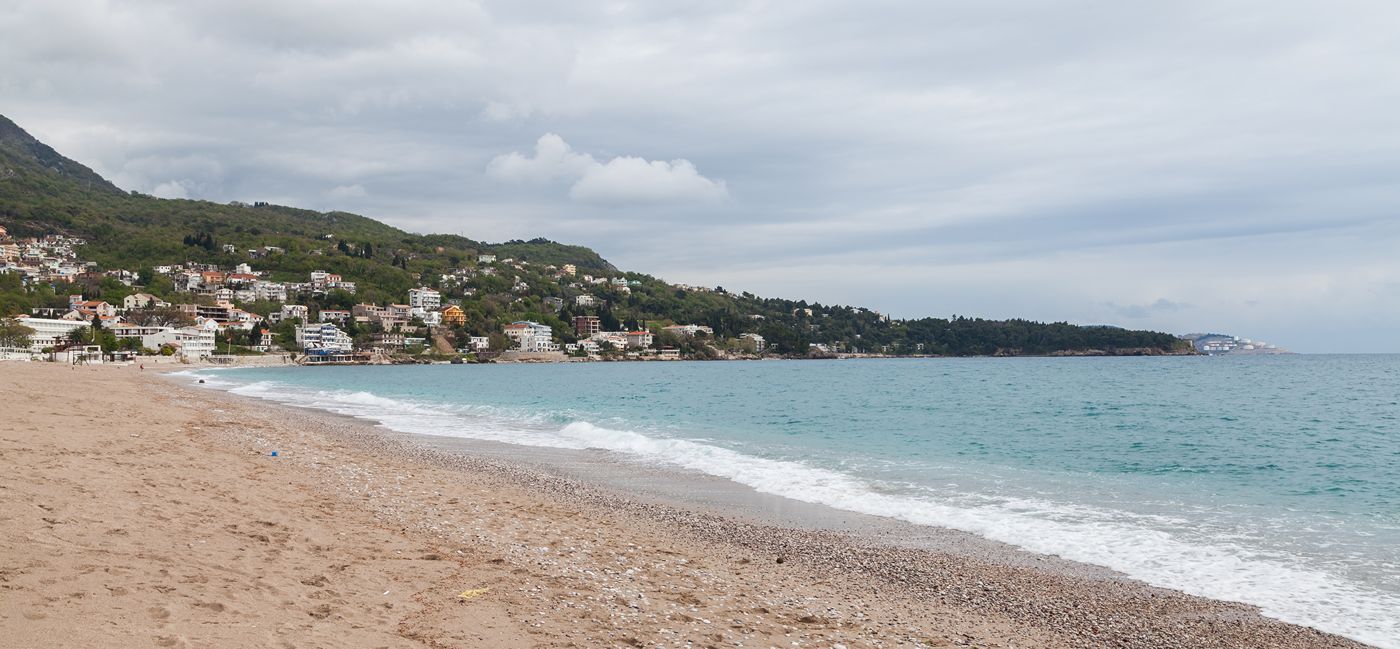 A wide view of the long sandy and pebble shore of Sutomore Beach with gentle waves on a cloudy day.