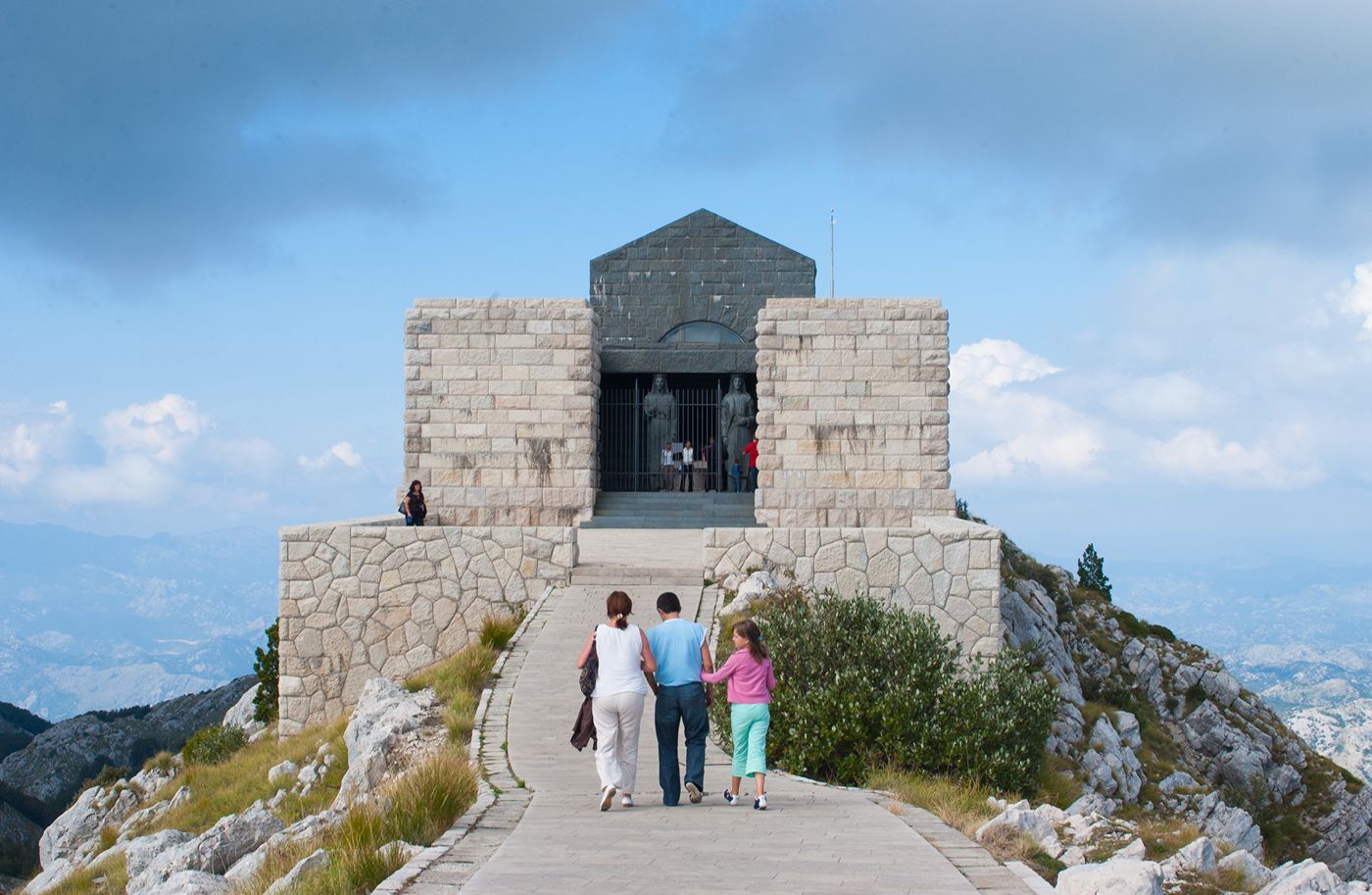 A family walking up the path to the entrance of the Njegoš Mausoleum, flanked by two giant stone statues.