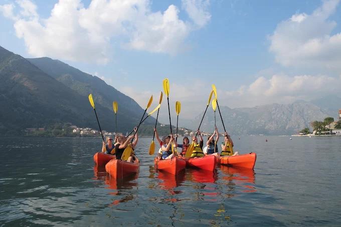 A happy group of kayakers raising their paddles in celebration on the calm waters of Boka Bay, with mountains surrounding them.