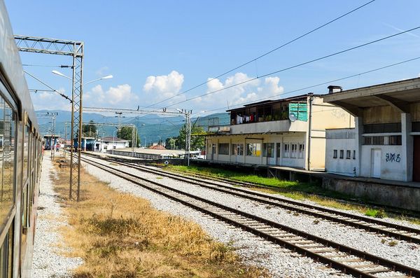 A view from a train window at the Bijelo Polje railway station in Montenegro, showing multiple tracks, weathered station buildings, and distant mountains under a blue sky.