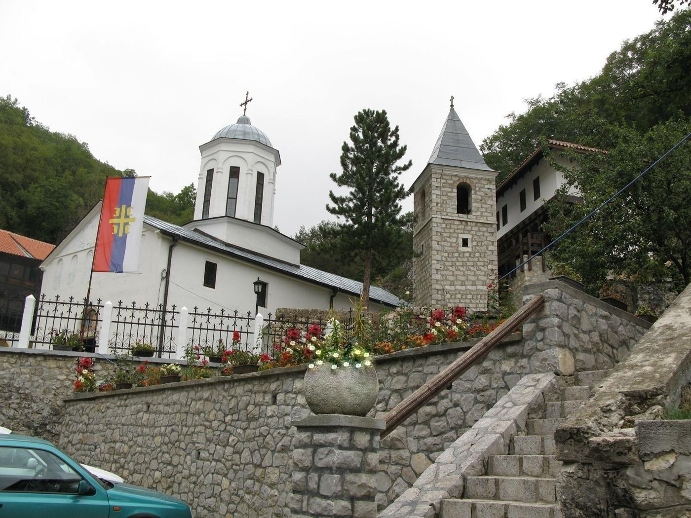 A view looking up a stone staircase towards the Holy Trinity Monastery complex, nestled on a wooded hillside.