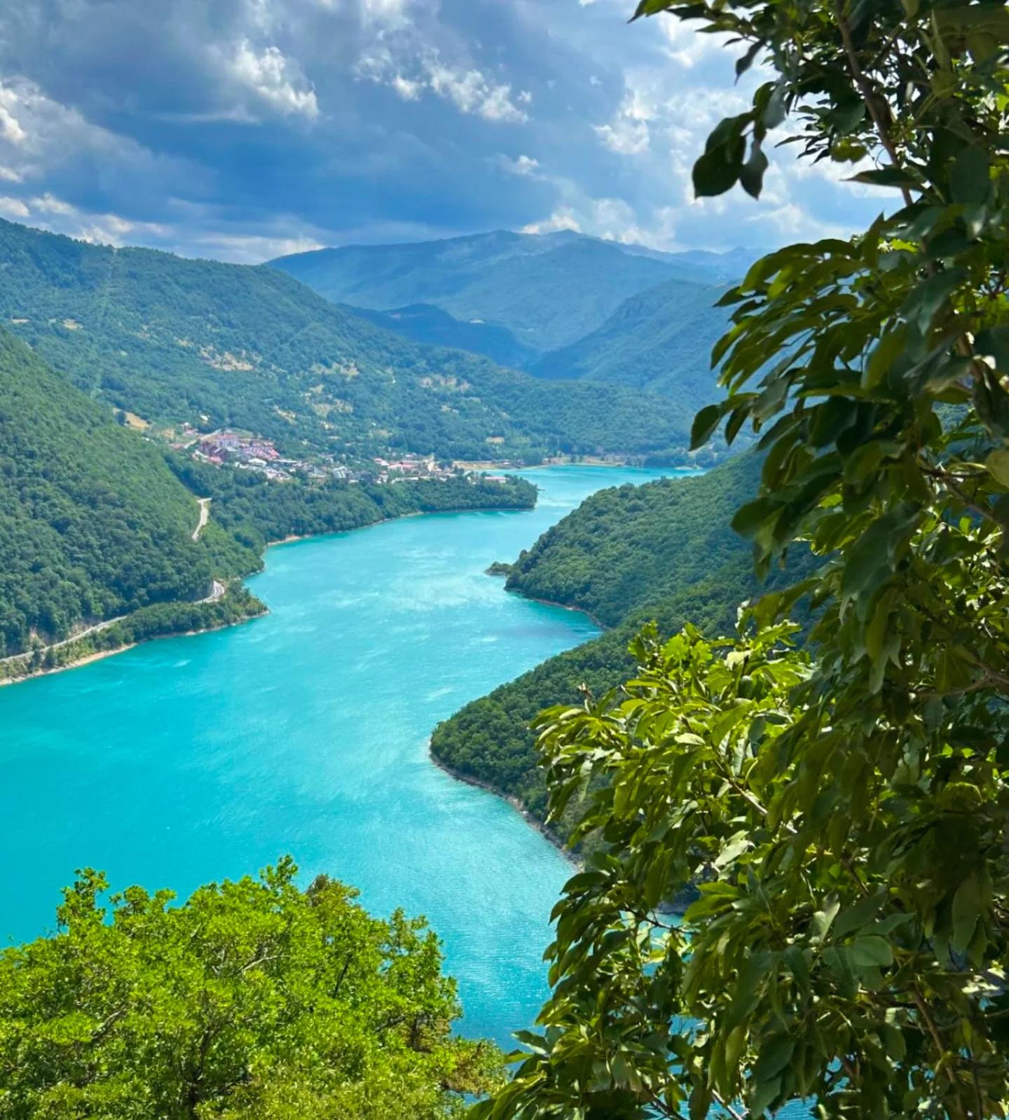 A stunning panoramic view of the bright turquoise Piva Lake winding through lush green mountains under a dramatic cloudy sky.