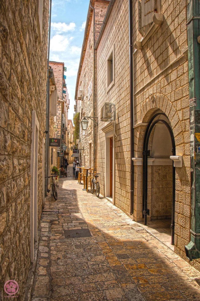 A sun-drenched narrow cobblestone street in Budva's Old Town, lined with historic stone buildings.