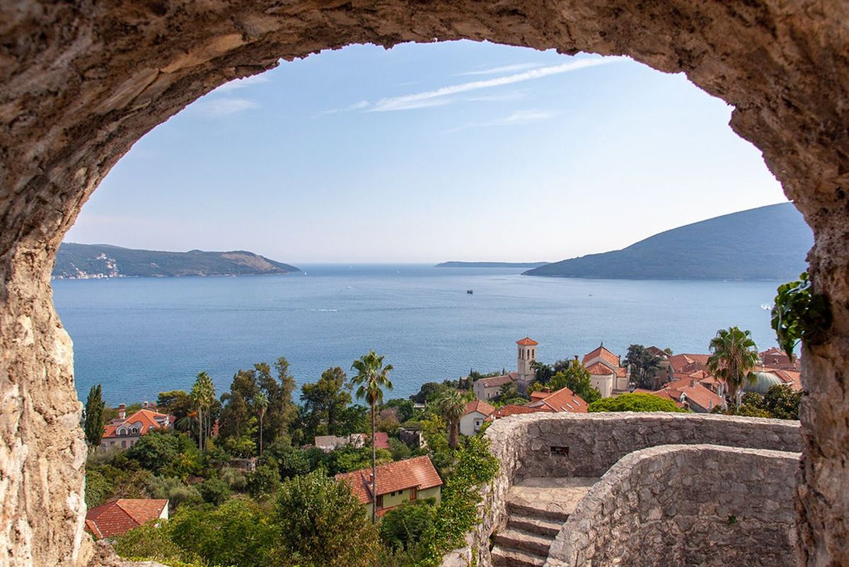 A scenic view of Herceg Novi and the entrance to the Bay of Kotor, framed by a rustic stone archway from a historic fortress