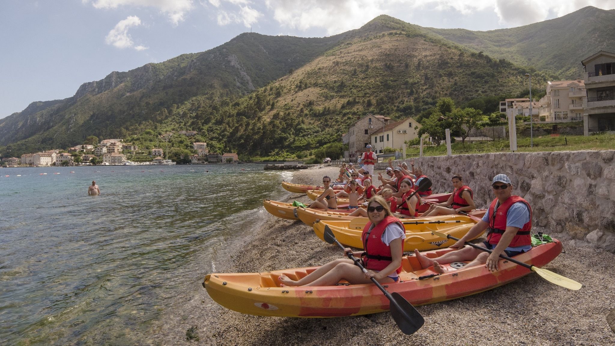 A group of tourists in yellow kayaks lined up on a pebble beach, preparing for a tour in the Bay of Kotor with mountains in the background.