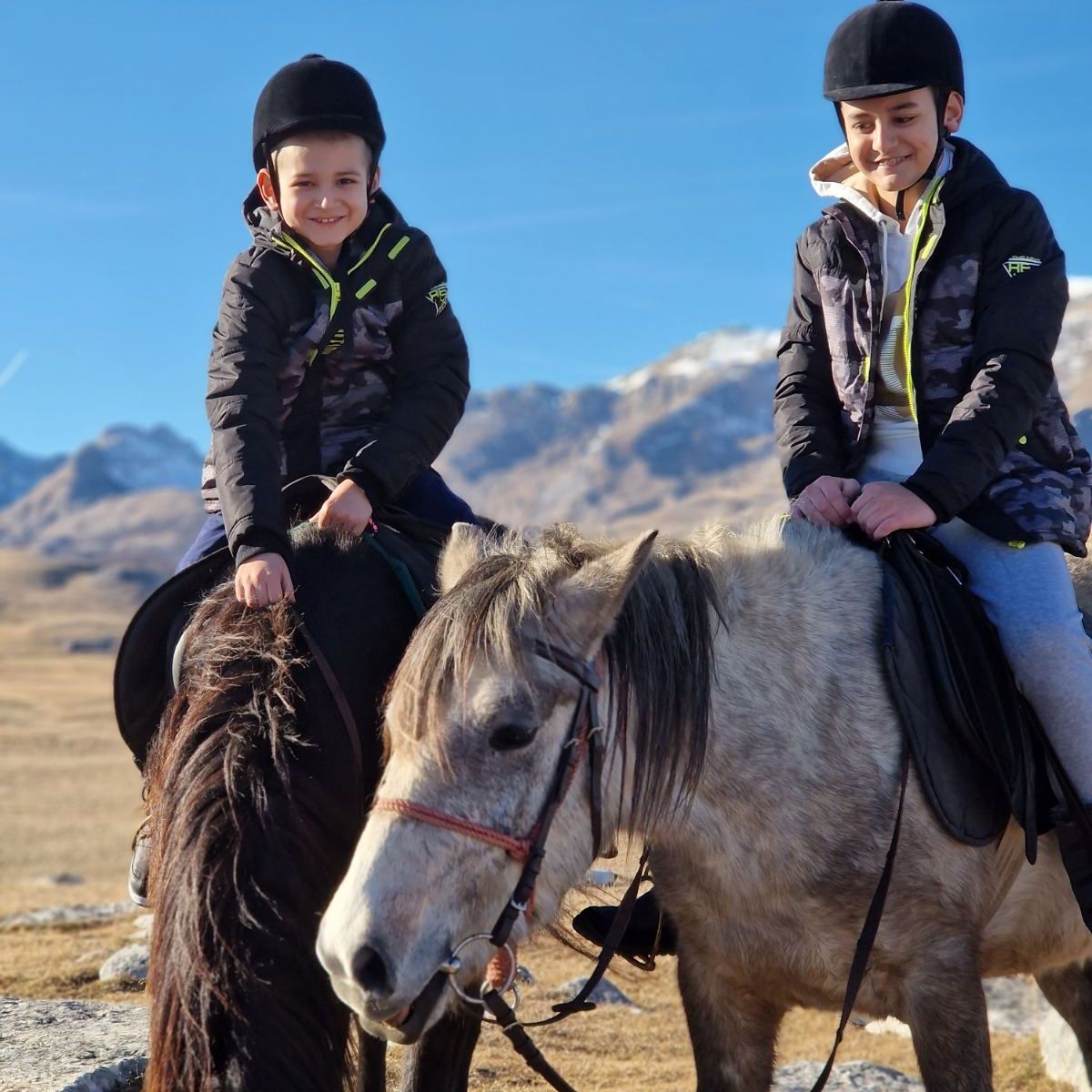Two smiling children wearing riding helmets and sitting on ponies in the Durmitor highlands, with snow-capped mountains in the background.