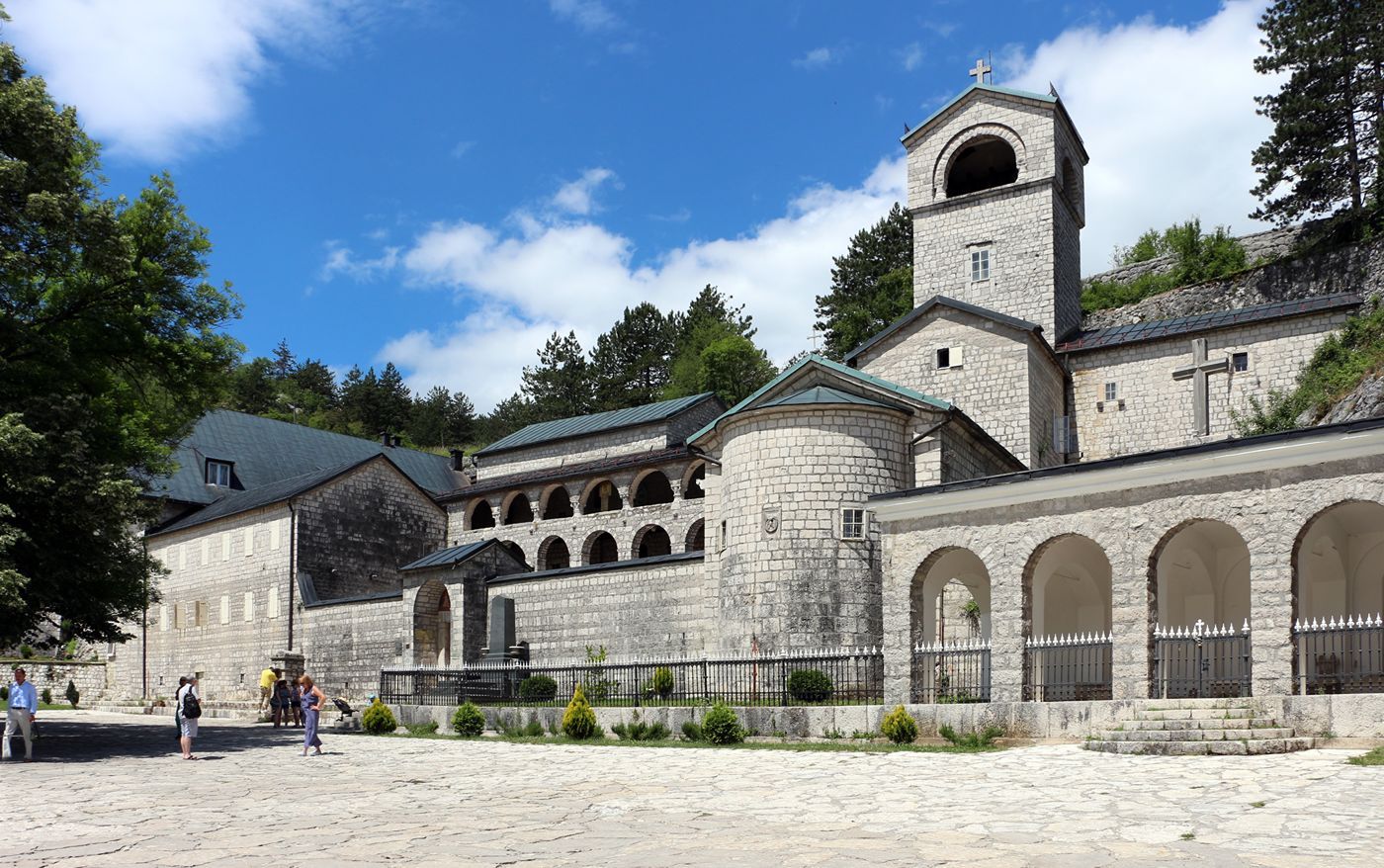 Visitors in the courtyard of the Cetinje Monastery on a sunny day, showing its stone arches and bell tower.