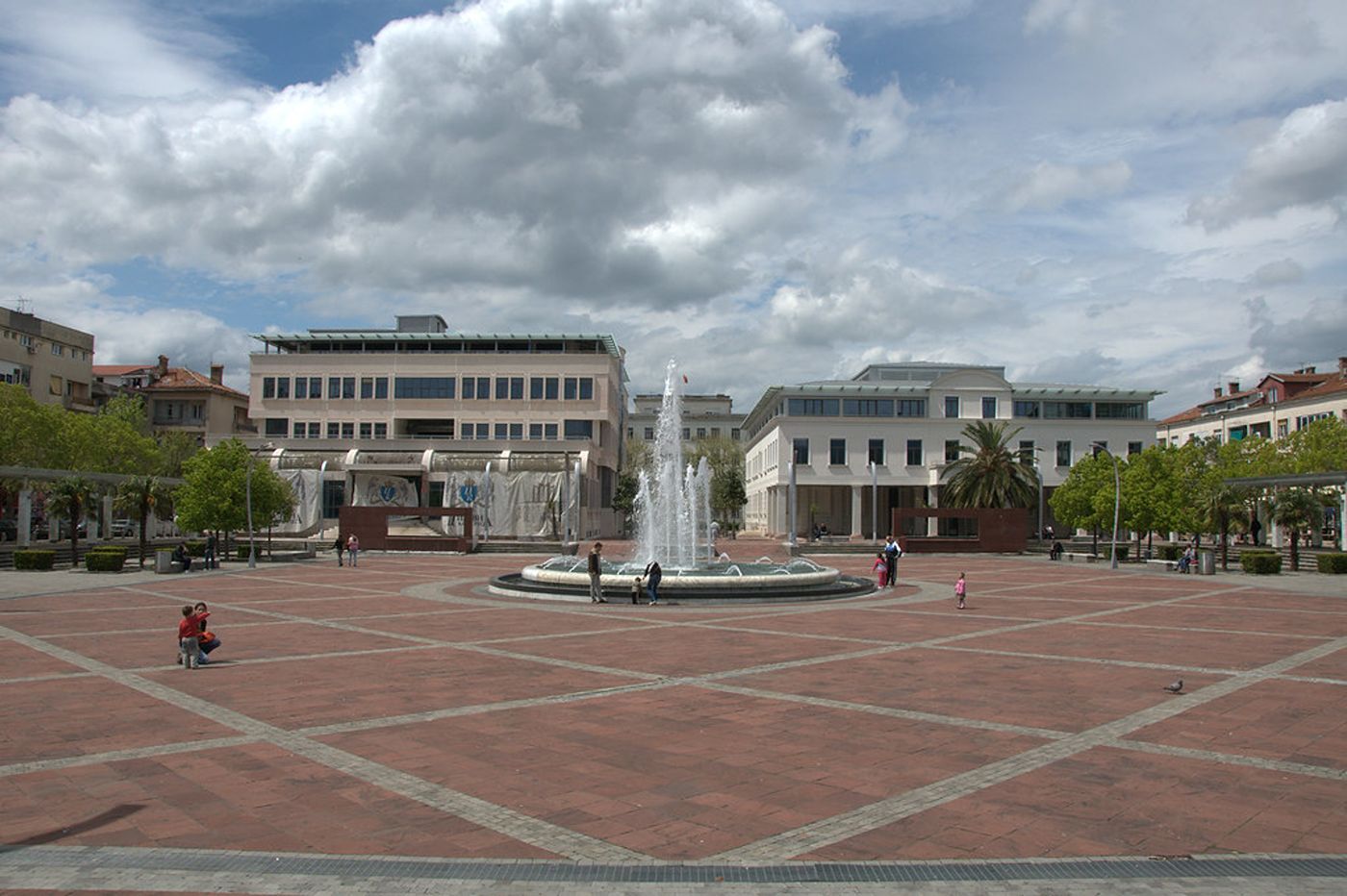 A wide view of Independence Square in Podgorica, with its central fountain and red tiled paving under a cloudy sky.