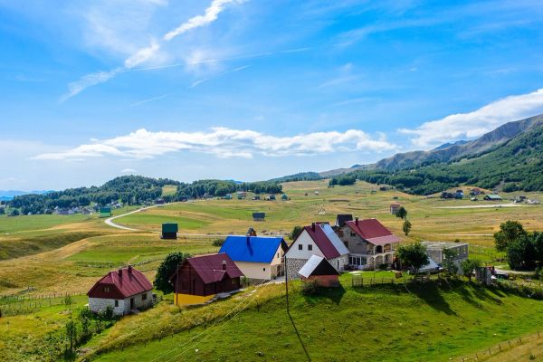 A scenic rural landscape near Žabljak, Montenegro, showing colorful houses scattered across vast, rolling green hills at the foot of the Durmitor mountains under a blue sky.