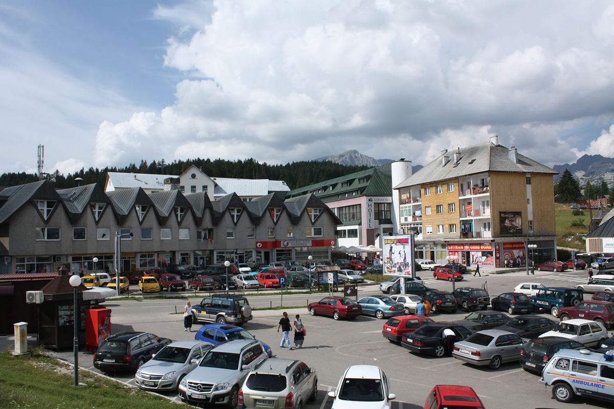 The town center of Žabljak, Montenegro, featuring alpine-style buildings and a bustling parking lot, with pine forests and a rocky Durmitor mountain peak in the background