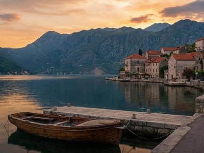 Scenic sunset view of the Bay of Kotor, Montenegro, featuring a traditional wooden boat tied to a stone pier in front of a historic waterfront village and mountains.