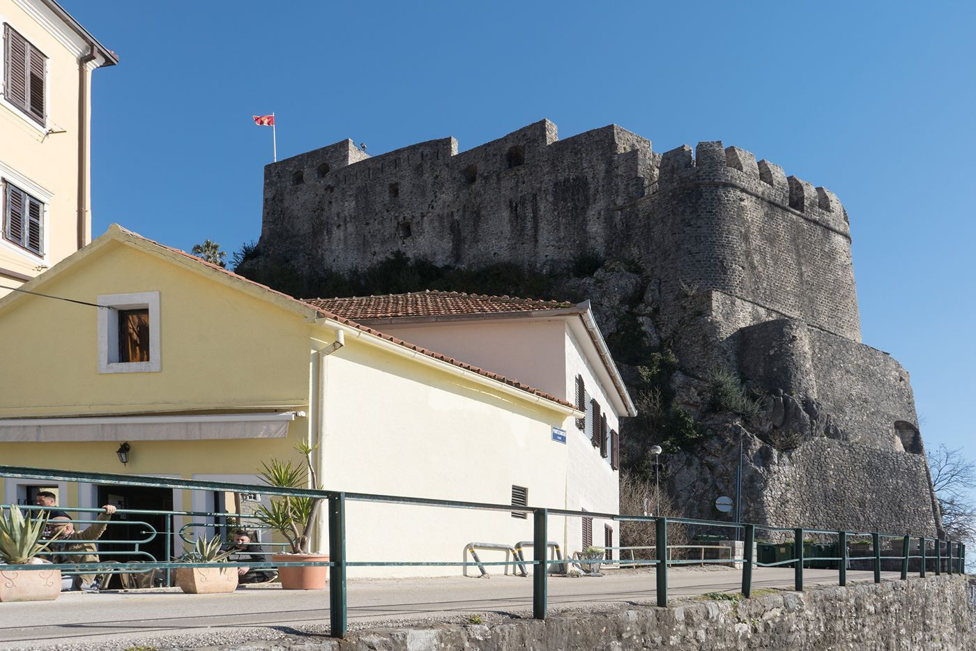 The imposing stone walls of the Forte Mare fortress towering above the houses in Herceg Novi against a clear blue sky.
