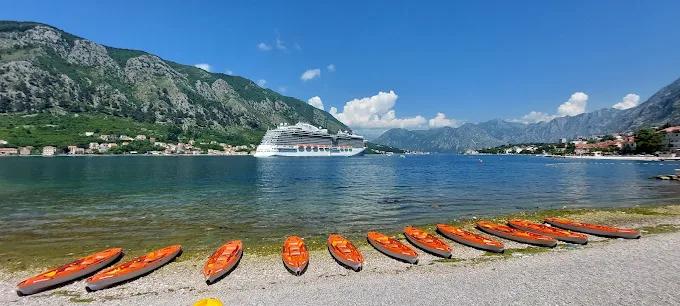 A line of orange kayaks resting on a pebble beach, with a large cruise ship and the mountains of Kotor Bay in the background.