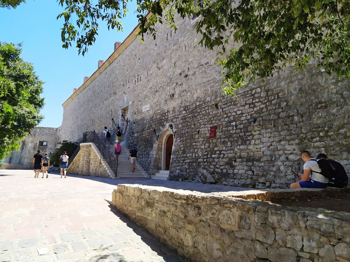 Tourists walking up the stone steps inside the sunny courtyard of the Budva Citadel.