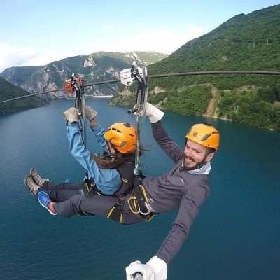 A smiling man and a woman taking a selfie while riding a tandem zipline high above the brilliant turquoise Piva Lake in Montenegro.