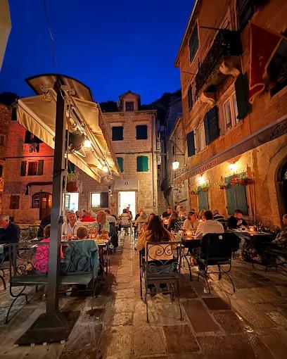 Outdoor tables of Konoba Scala Santa full of customers dining at night in a charming, historic square in Kotor.