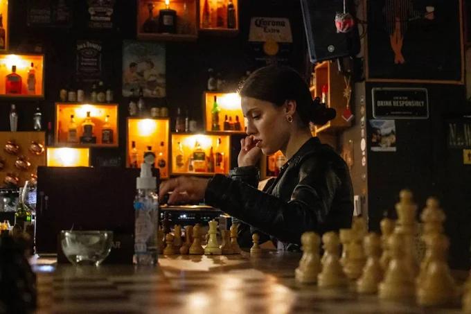 A woman in a leather jacket concentrates on a game of chess at the bar, with the warm glow of the liquor shelves in the background.