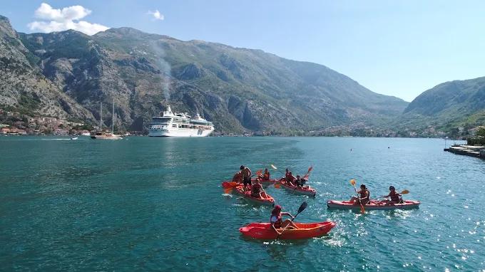 A group of kayakers paddling in Boka Bay, with a large cruise ship and the historic town of Kotor visible across the water.