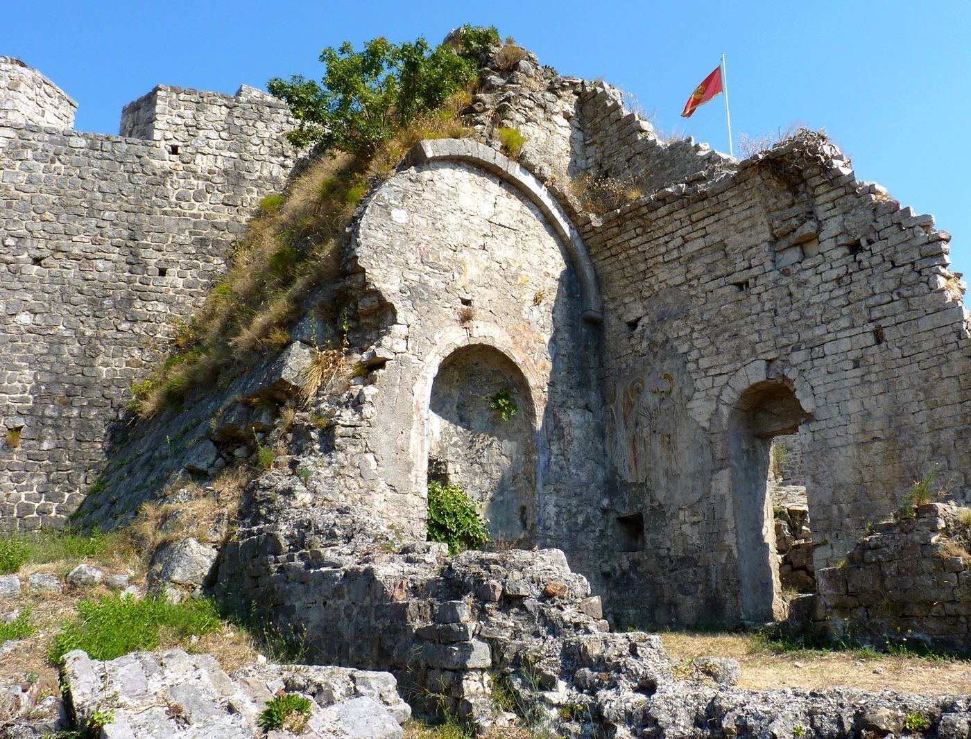 The crumbling stone apse of a ruined church within the historic walls of Stari Bar, with a Montenegrin flag.