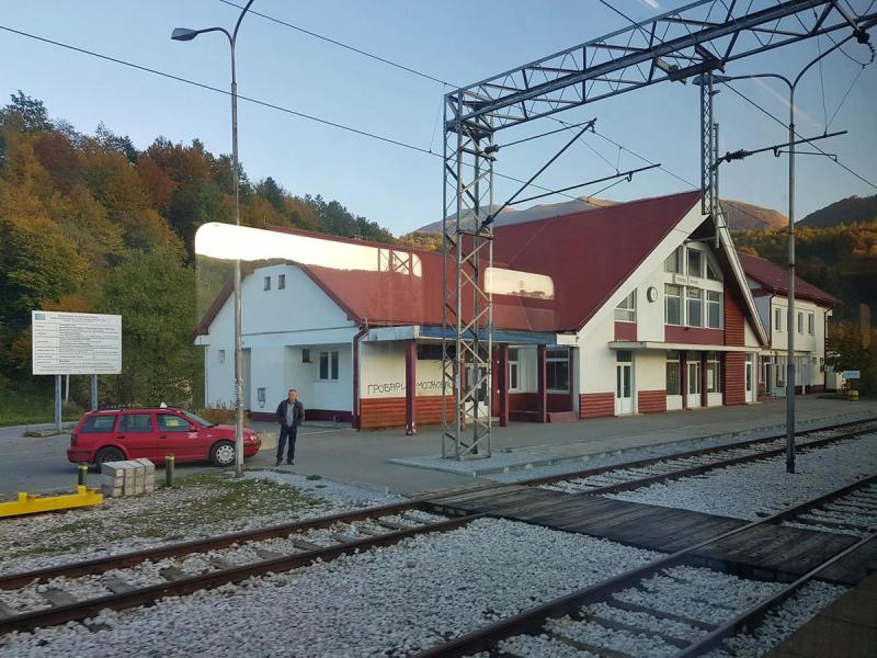 A view from a train of the Mojkovac railway station in Montenegro, showing the station building with a red roof, a car parked outside, and autumn-colored hills in the background