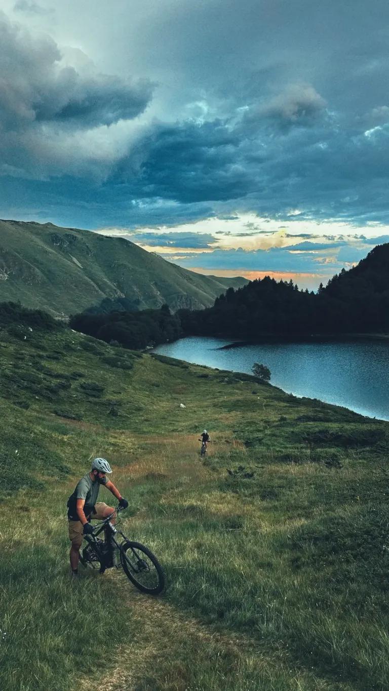 Two mountain bikers on a grassy trail overlooking a serene mountain lake under a dramatic sunset sky.