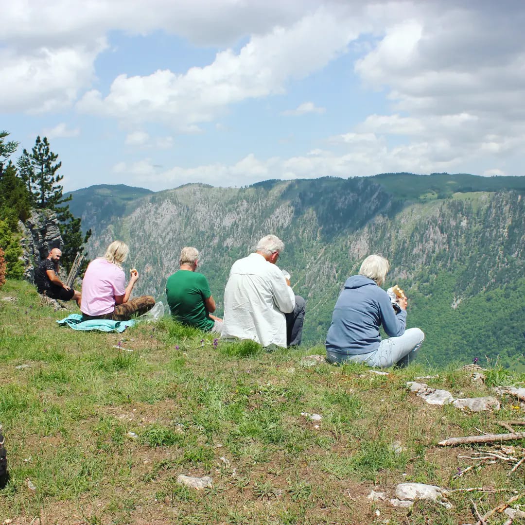 Grupa turystów robiąca sobie przerwę, siedząca na trawiastym klifie i jedząca, z widokiem na rozległy, zalesiony kanion pod częściowo zachmurzonym niebem.