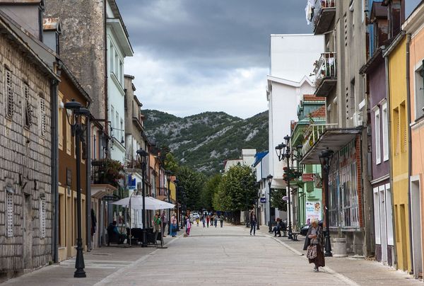 A view down the main pedestrian street of Cetinje, Montenegro, lined with historic buildings and cafes, with people strolling under a cloudy sky and a karst mountain in the background.