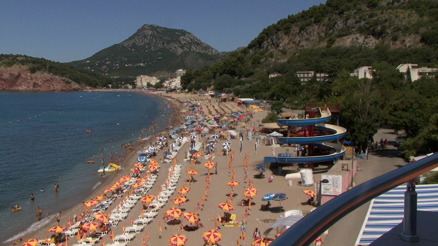 A crowded Sutomore Beach in summer, packed with colorful umbrellas and sunbathers, with a waterslide nearby.