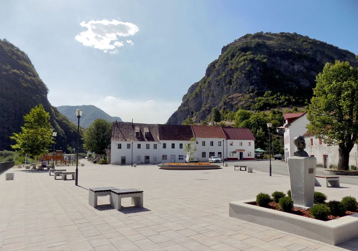 The spacious main square of Šavnik, Montenegro, a small town nestled in a deep valley, showing traditional white buildings, a monument, and steep green mountains rising on all sides.