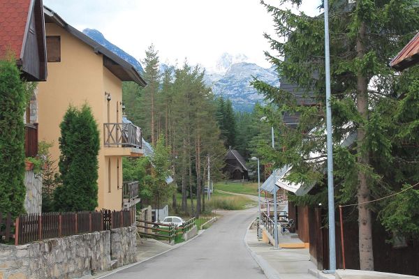 A quiet street in Žabljak, Montenegro, lined with alpine-style chalets and tall pine trees, with a view of a snow-dusted Durmitor mountain peak in the distance