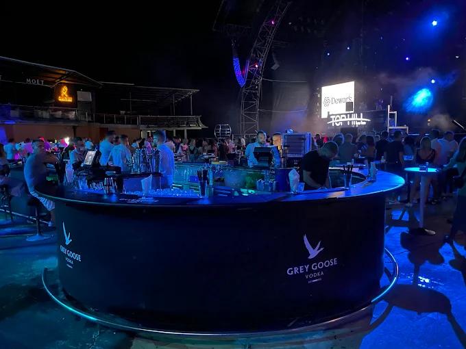 Top Hill 夜总会一个繁忙的圆形吧台，调酒师在蓝色和紫色的派对灯光下为顾客准备饮品。