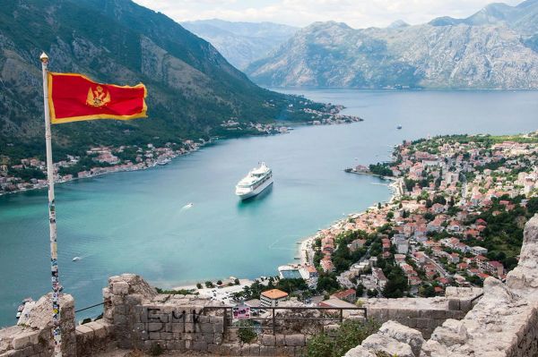 The flag of Montenegro flying on a flagpole from the Kotor fortress walls, with a panoramic view of the Bay of Kotor, a cruise ship, and the surrounding mountains.