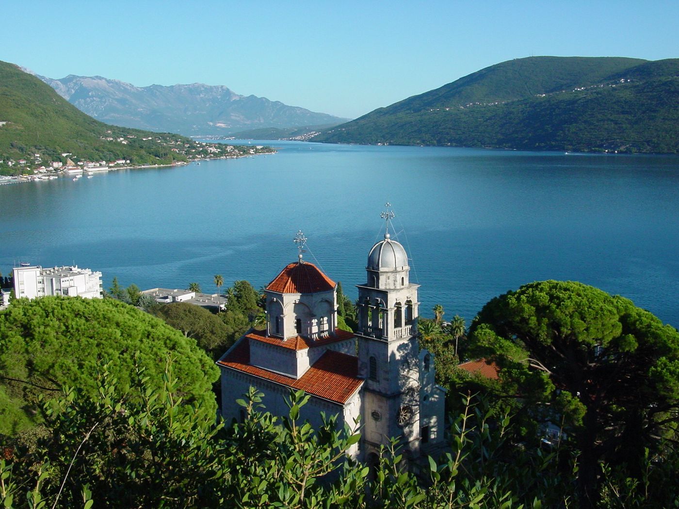 A scenic view of the Savina Monastery and its bell tower, with the tranquil Bay of Kotor and mountains in the distance.