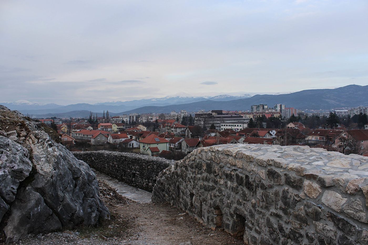  A view from the stone walls of the Bedem Fortress overlooking the city of Nikšić, Montenegro, with distant snow-capped mountains under a cloudy sky