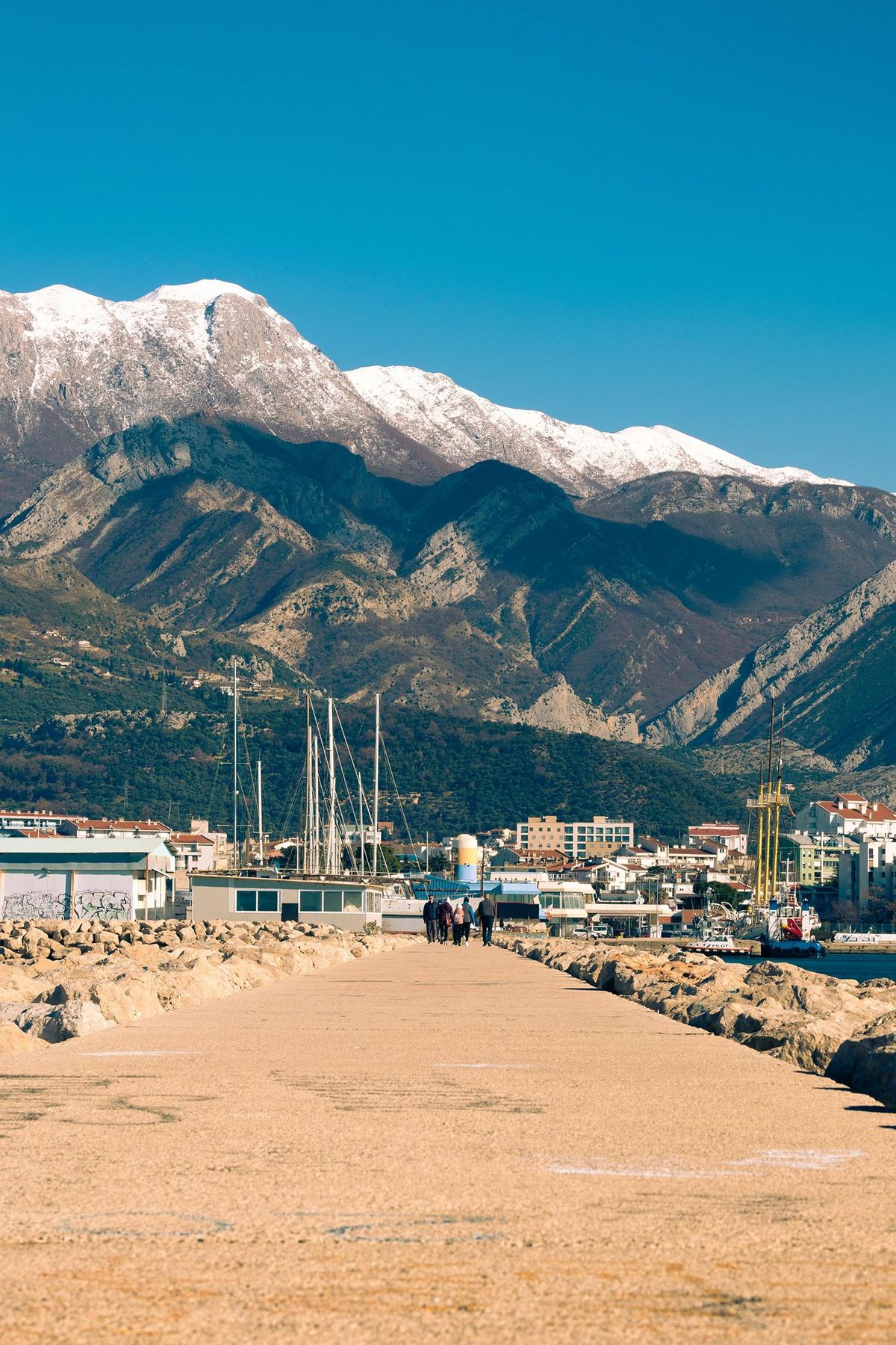 A view down the stone pier of the Port of Bar, Montenegro, with the city and towering, snow-capped mountains in the background under a clear blue sky