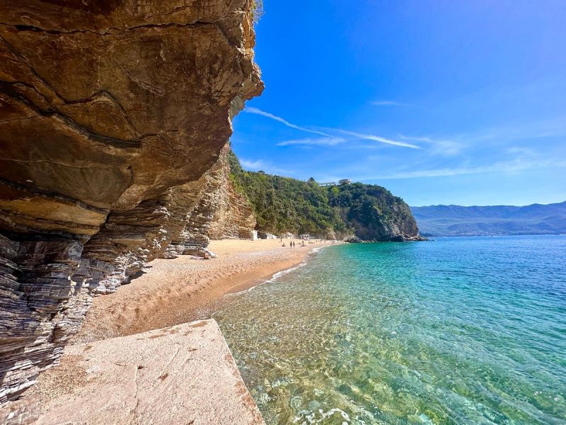 The crystal-clear turquoise water and sandy shore of Mogren Beach, framed by unique layered rock cliffs.