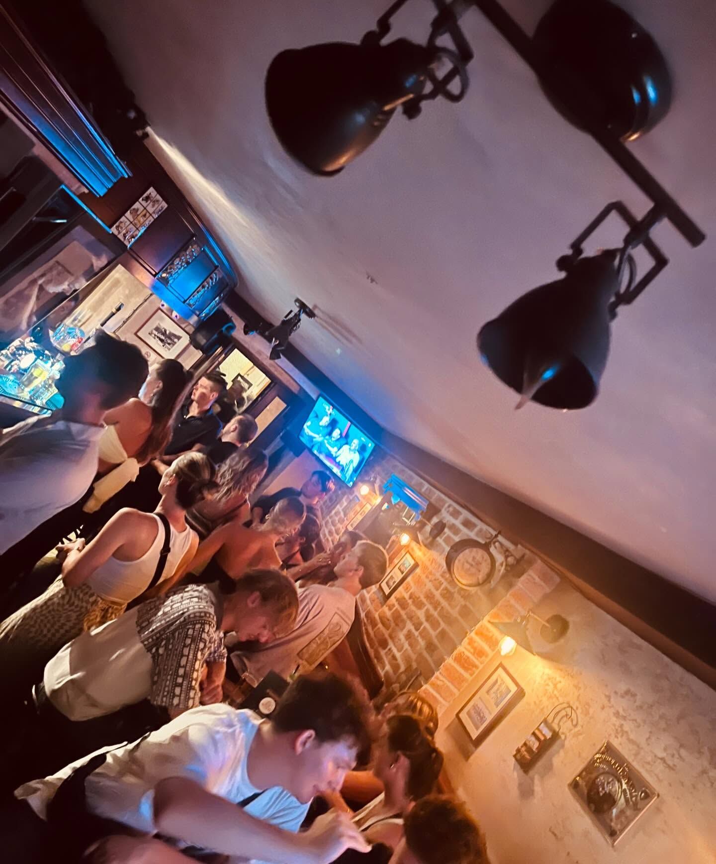The lively interior of the pub, with a crowd of people gathered around the bar in a warm, atmospheric setting with brick walls.