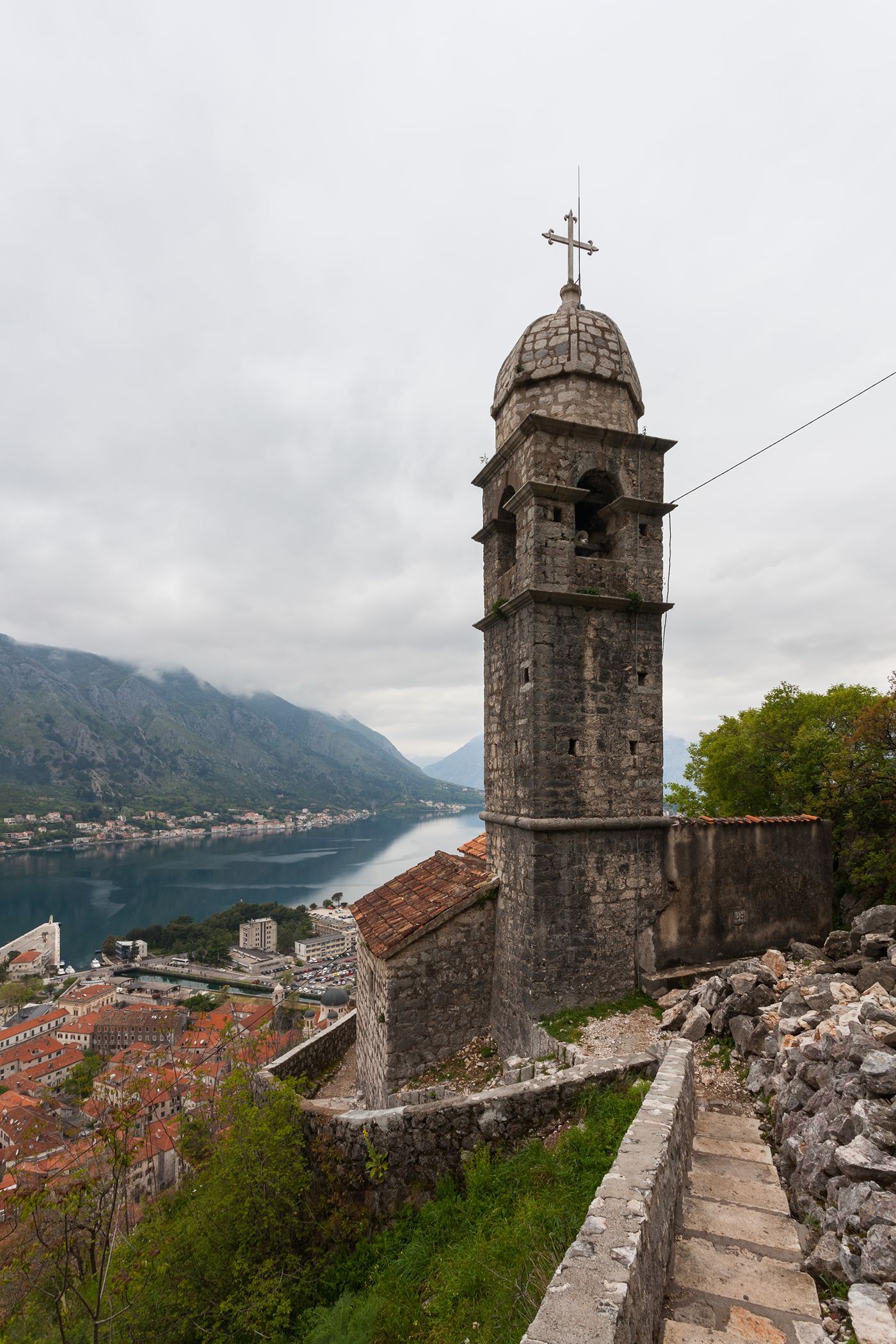 The stone bell tower of the Church of Our Lady of Remedy overlooking the Bay of Kotor on a cloudy day.