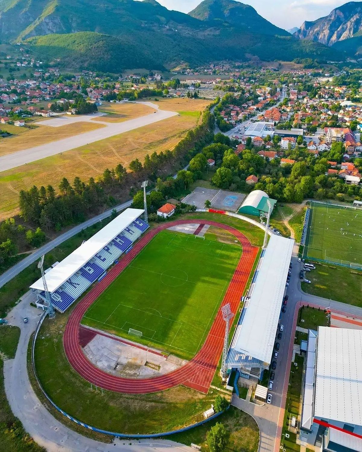 Pogled iz vazduha na Gradski stadion u Beranama, Crna Gora, koji prikazuje fudbalski teren, crvenu atletsku stazu i tribine, sa malom aerodromskom pistom i gradom smještenim u planinskoj dolini