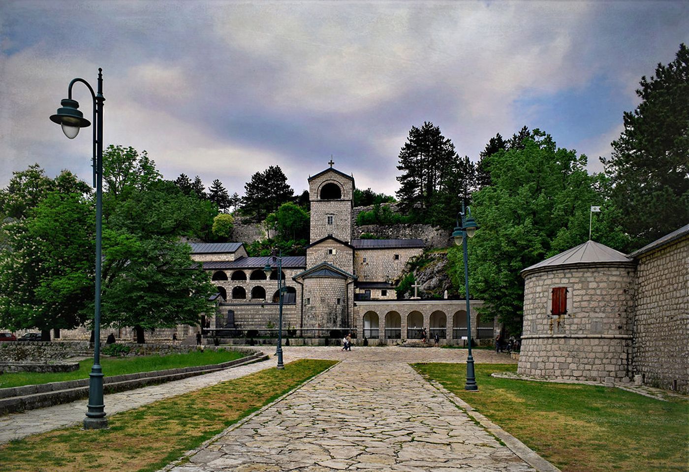 The historic stone complex of the Cetinje Monastery at the foot of a hill, with a path leading towards it.