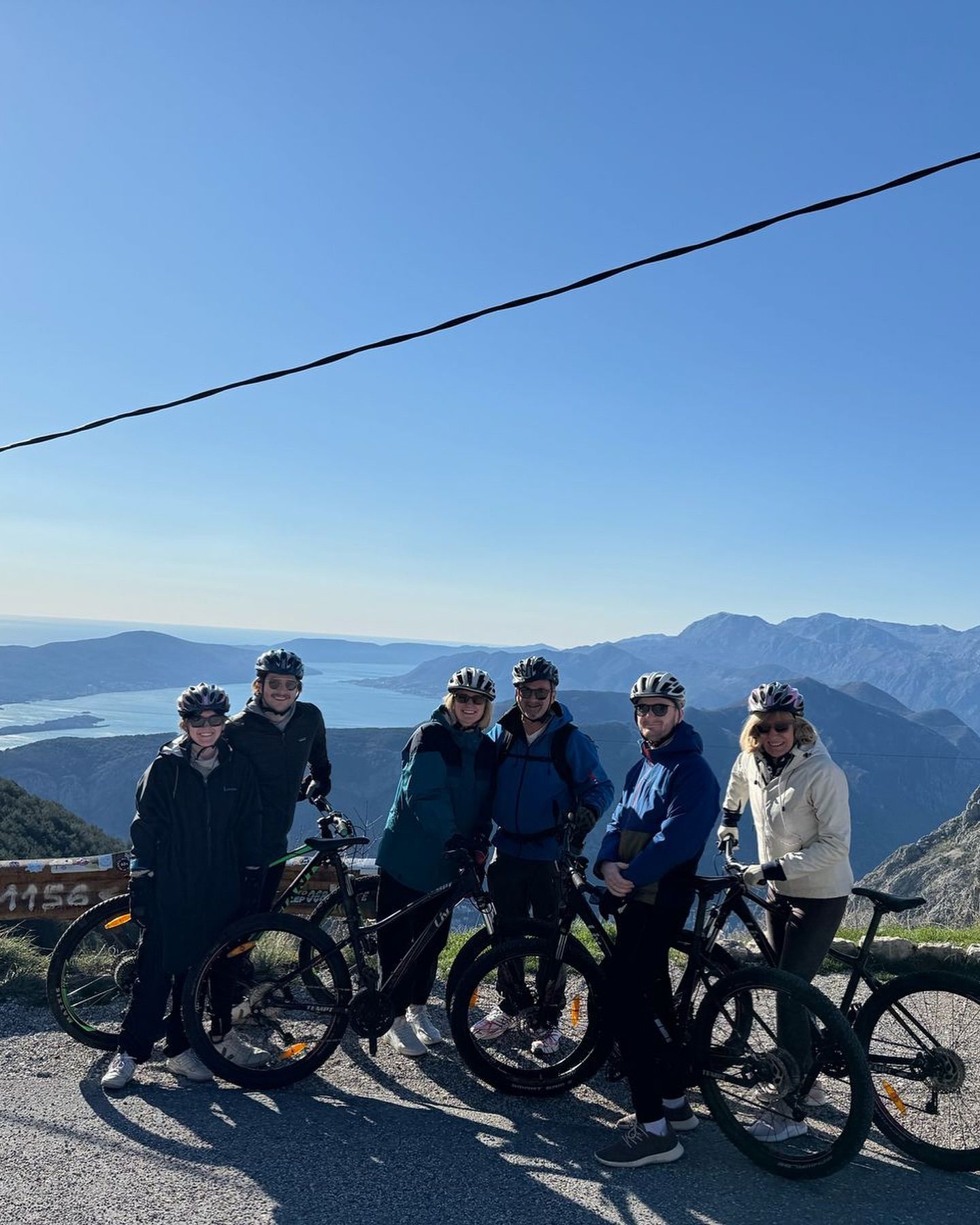 Grupa nasmijanih biciklista sa kacigama pozira sa svojim brdskim biciklima na putu sa pogledom na prelijepi Bokokotorski zaliv u Crnoj Gori.
