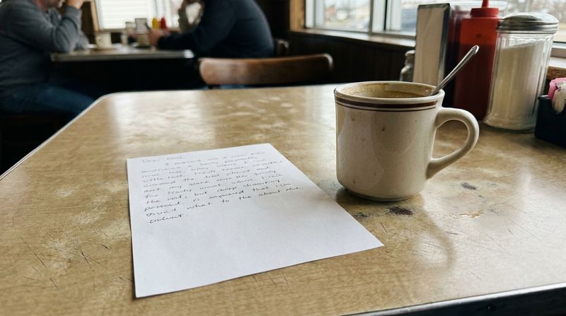 A handwritten note on plain paper resting on a diner table beside a coffee cup
