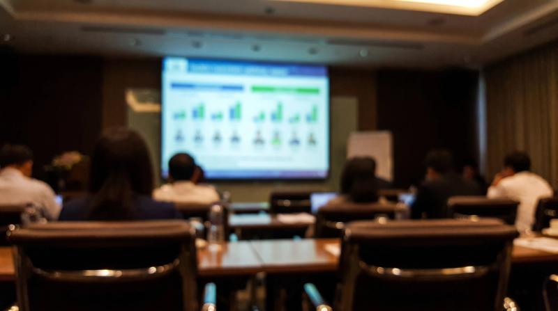A blurred conference room presentation screen showing rows of figures