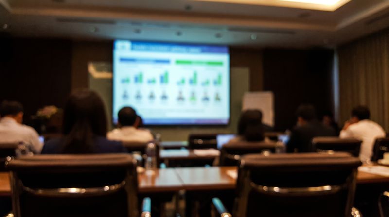 A blurred conference room presentation screen showing rows of figures