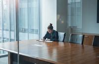 A young woman sitting alone at the end of a large glass-walled corporate conference room with a notepad