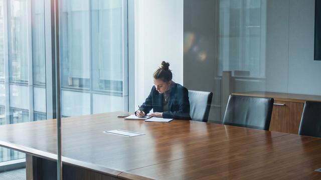 A young woman sitting alone at the end of a large glass-walled corporate conference room with a notepad