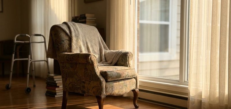 An empty armchair by a bright window in a care home, soft afternoon light