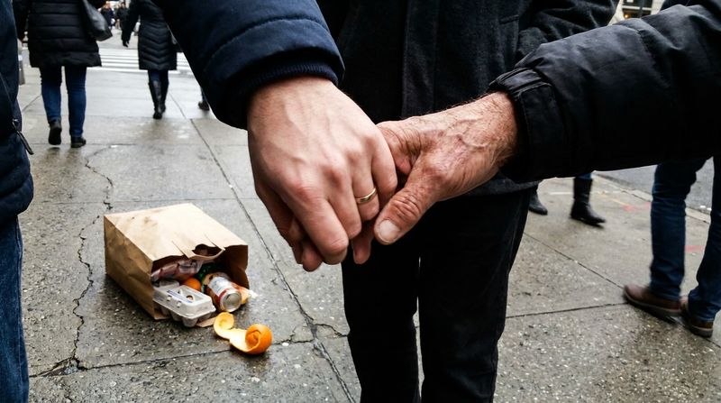 A young mans hand tightly holding an older mans hand on a city sidewalk, a split grocery bag nearby